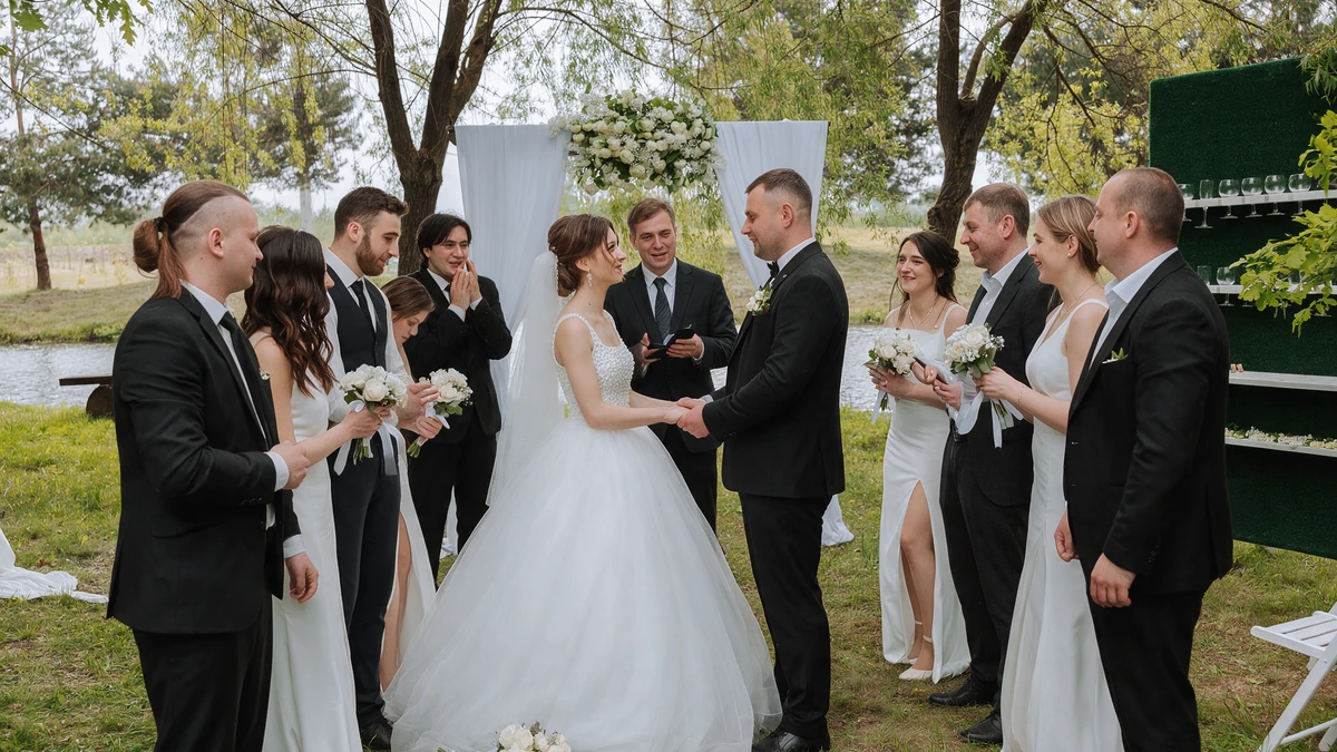 Couple holding hands at an outdoor ceremony as their officiant reads, with the wedding party gathered in a semicircle around them