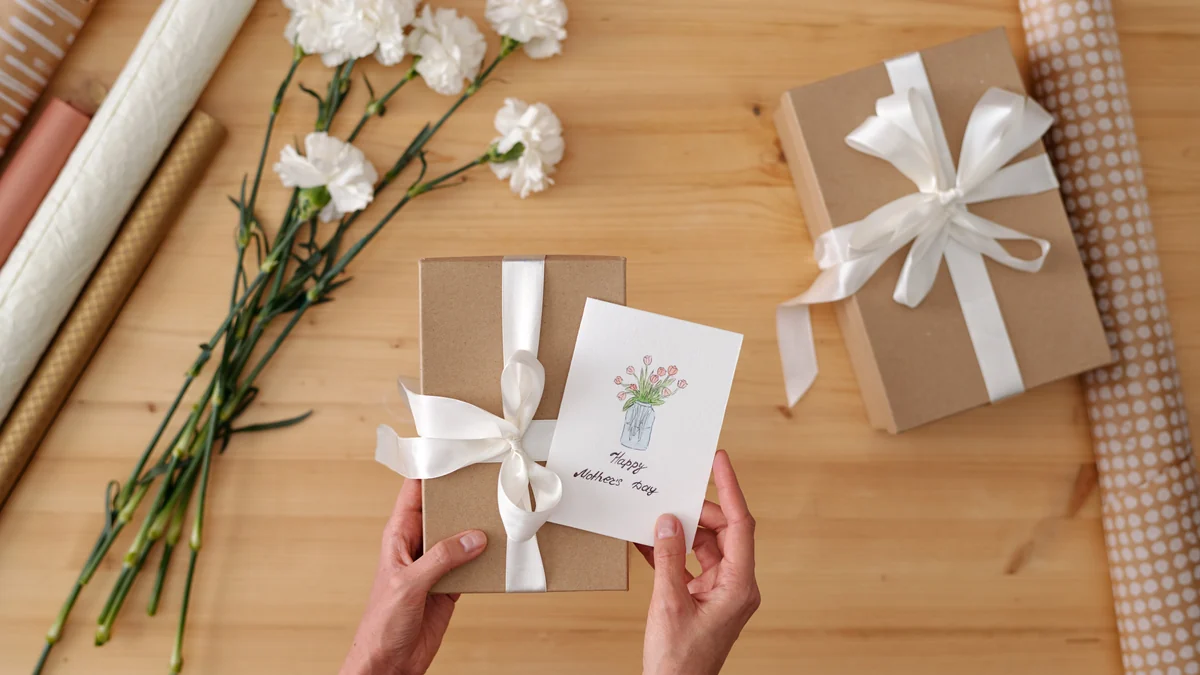 Hands holding a kraft paper wrapped gift with a ribbon and a small handwritten card beside white carnations