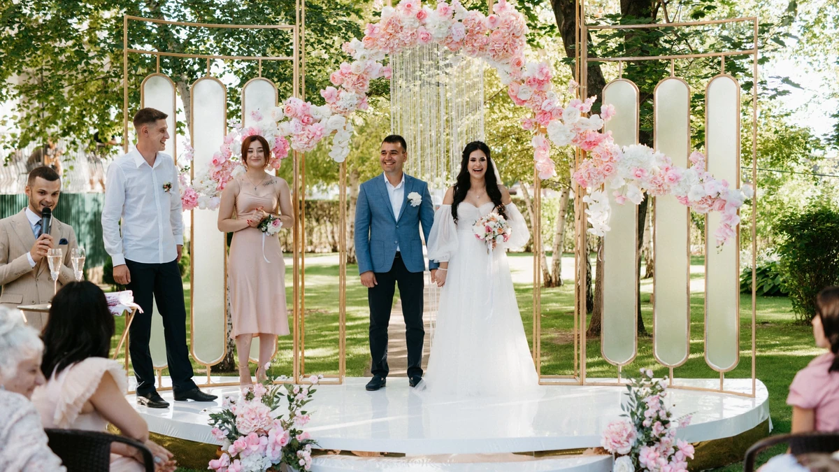 Couple standing before a gold frame arch dressed with pink and white flowers at an outdoor ceremony as guests look on