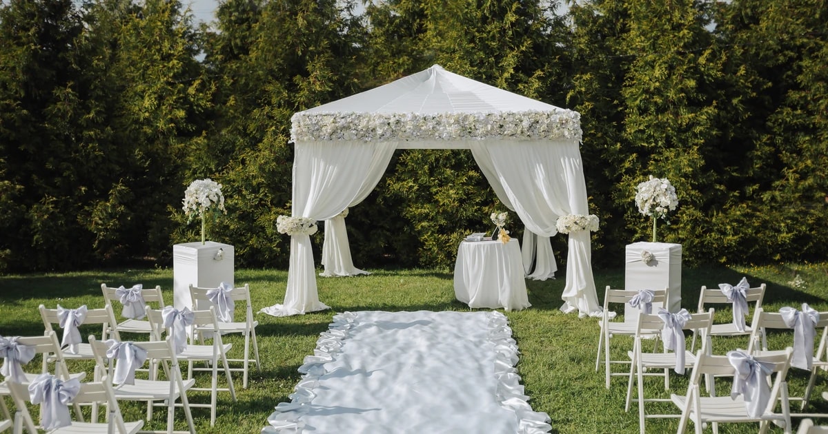 Outdoor wedding ceremony set up with white chairs lining a petal-edged aisle leading to a floral draped marquee