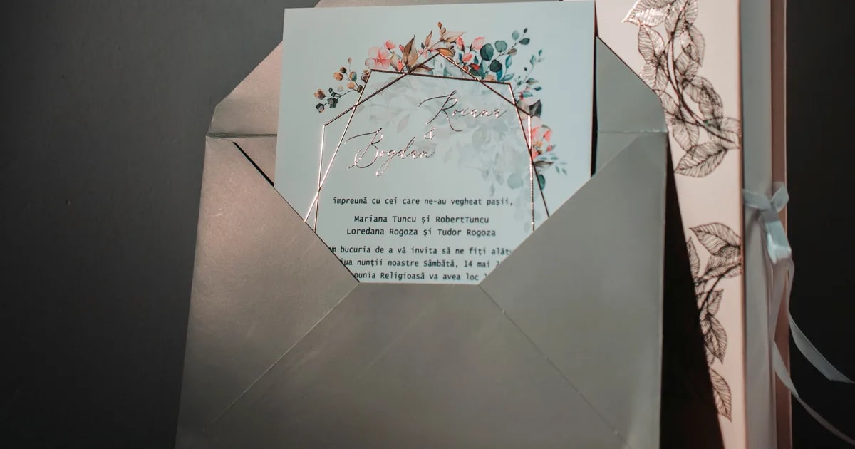 Sage green wedding invitation with a rose gold geometric frame and watercolour florals, tucked into a matching envelope beside a foil-stamped keepsake box, an example of a complete wedding invitation suite