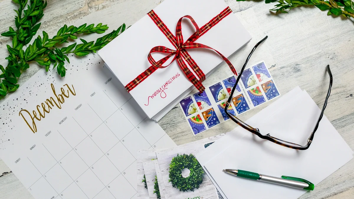 Monthly calendar laid beside a ribbon tied bundle of cards, a sheet of stamps and a pen on a white wood desk