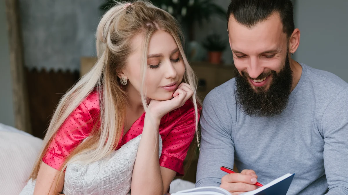 Couple sitting together on a sofa, one writing in a notebook while the other watches