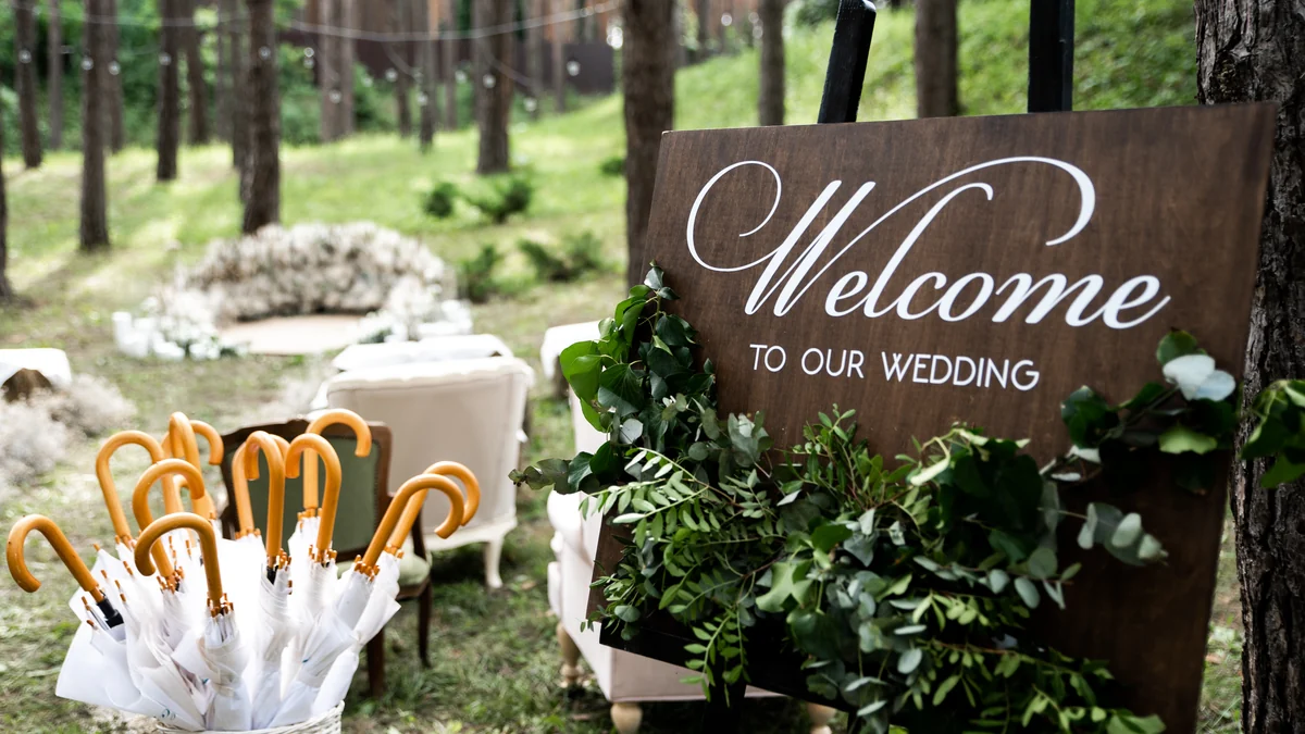 Wooden welcome sign with calligraphy and greenery beside a basket of umbrellas at an outdoor ceremony